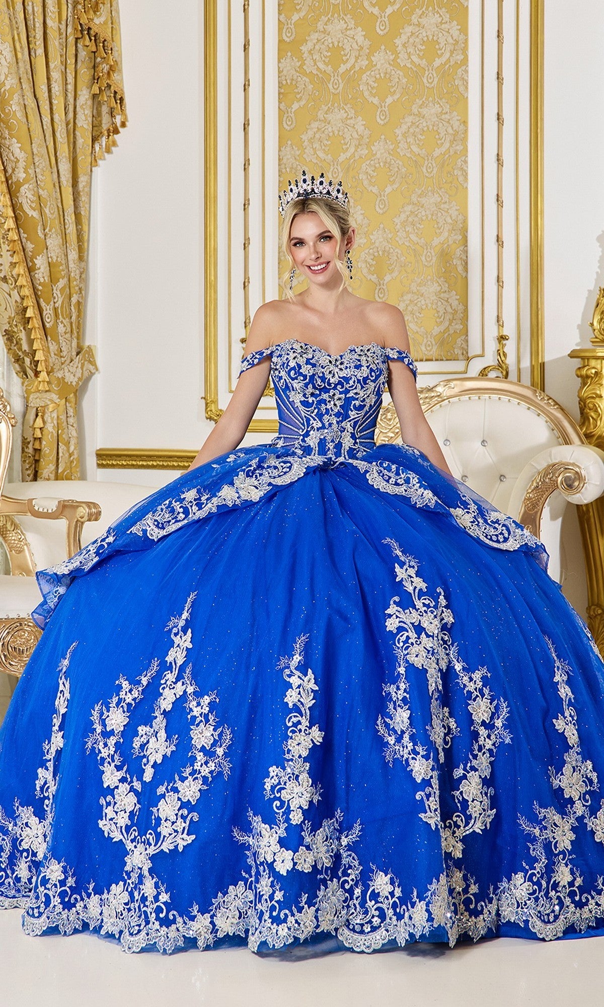 A woman in the Quinceanera Dress 5142 by Dancing Queen, royal blue with a corset bodice and floral embroidery, sits on a white and gold sofa with a tiara in an ornately decorated room.